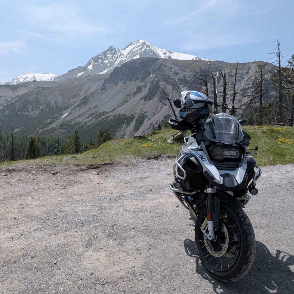 Grau-schwarze BMW R 1200 GS Adventure steht auf Schotter mit Helm am Lenker, vor einem verschneiten Berggipfel.