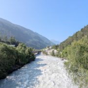 Reißender, milchig-weißer Fluss in einem grünen Alpental, mit Wäldern an den Hängen und einer niedrigen Brücke im Hintergrund.