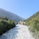 Reißender, milchig-weißer Fluss in einem grünen Alpental, mit Wäldern an den Hängen und einer niedrigen Brücke im Hintergrund.