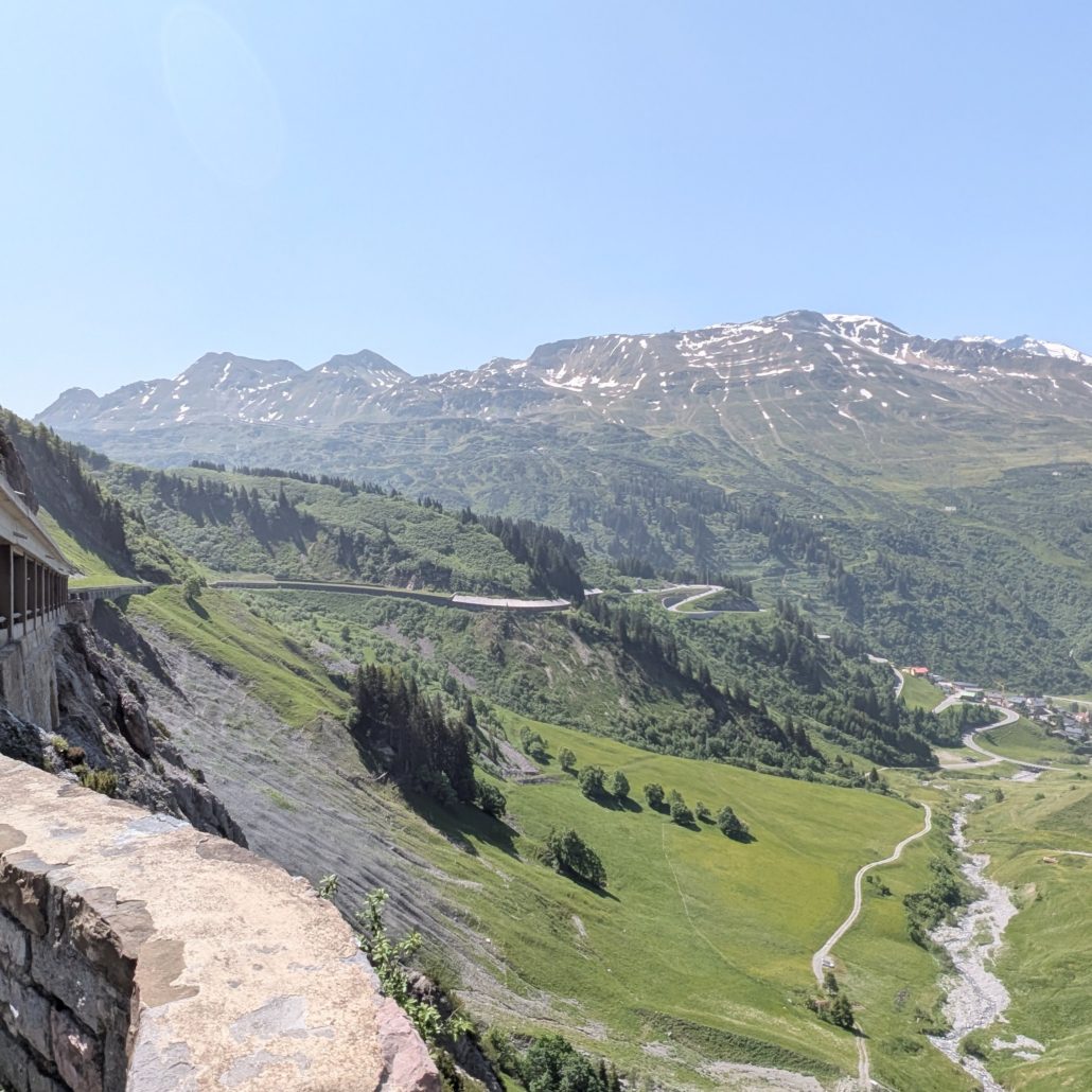 Majestätische Alpenlandschaft mit grünen Berghängen, einer Serpentinenstraße und schneebedeckten Gipfeln unter blauem Himmel.