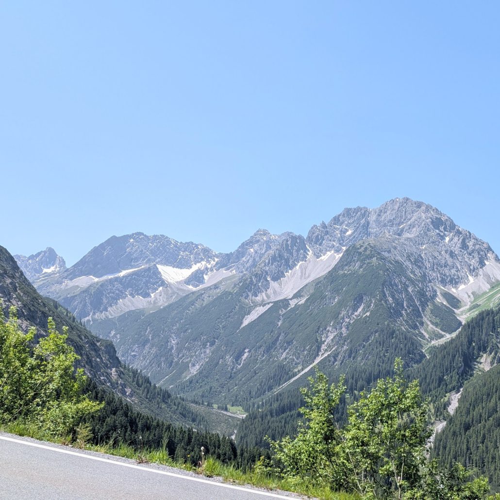 Asphaltierte Straße und bewaldetes Gebirgstal mit hohen Gipfeln, teilweise schneebedeckt, unter klarem, hellblauem Himmel.