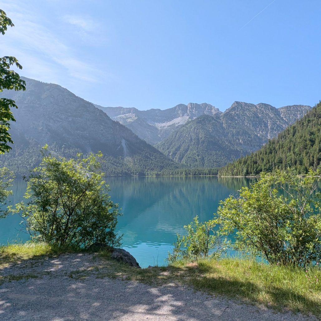 Kiesweg und grünes Gebüsch im Vordergrund, dahinter der türkisfarbene Blindsee in Österreich, umrahmt von bewaldeten Bergen.