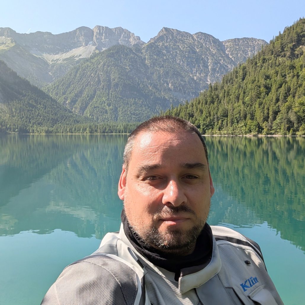 Mann im Selfie-Format vor einem Bergsee mit türkisblauem Wasser, das die bewaldeten Berghänge spiegelt.