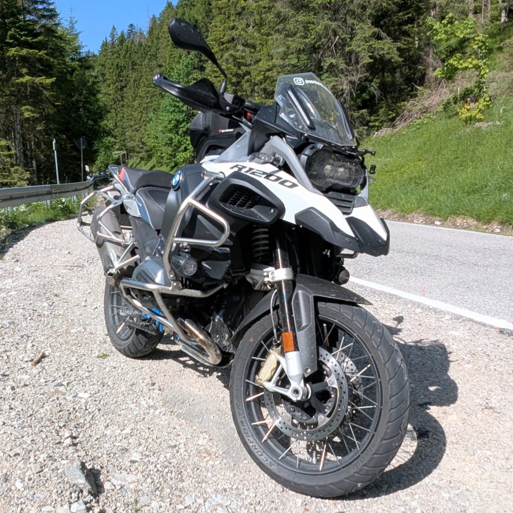Grau-weißes BMW R 1200 GS Motorrad steht am Rand einer kurvigen Straße neben einem schottrigen Seitenstreifen in einem Waldgebiet.