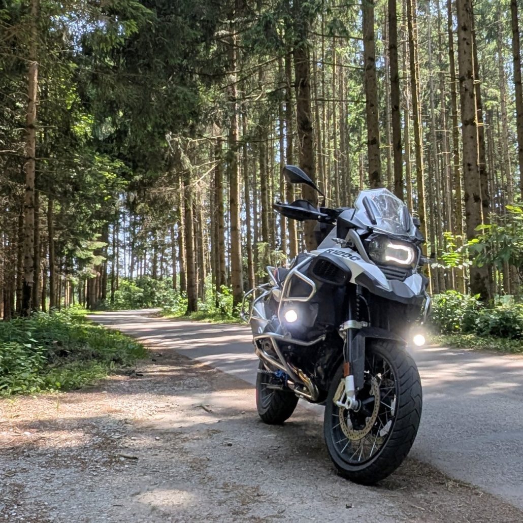 Grau-weißes BMW R 1200 GS Motorrad mit leuchtendem LED-Scheinwerfer auf einer asphaltierten Waldstraße, umgeben von hohen Nadelbäumen.