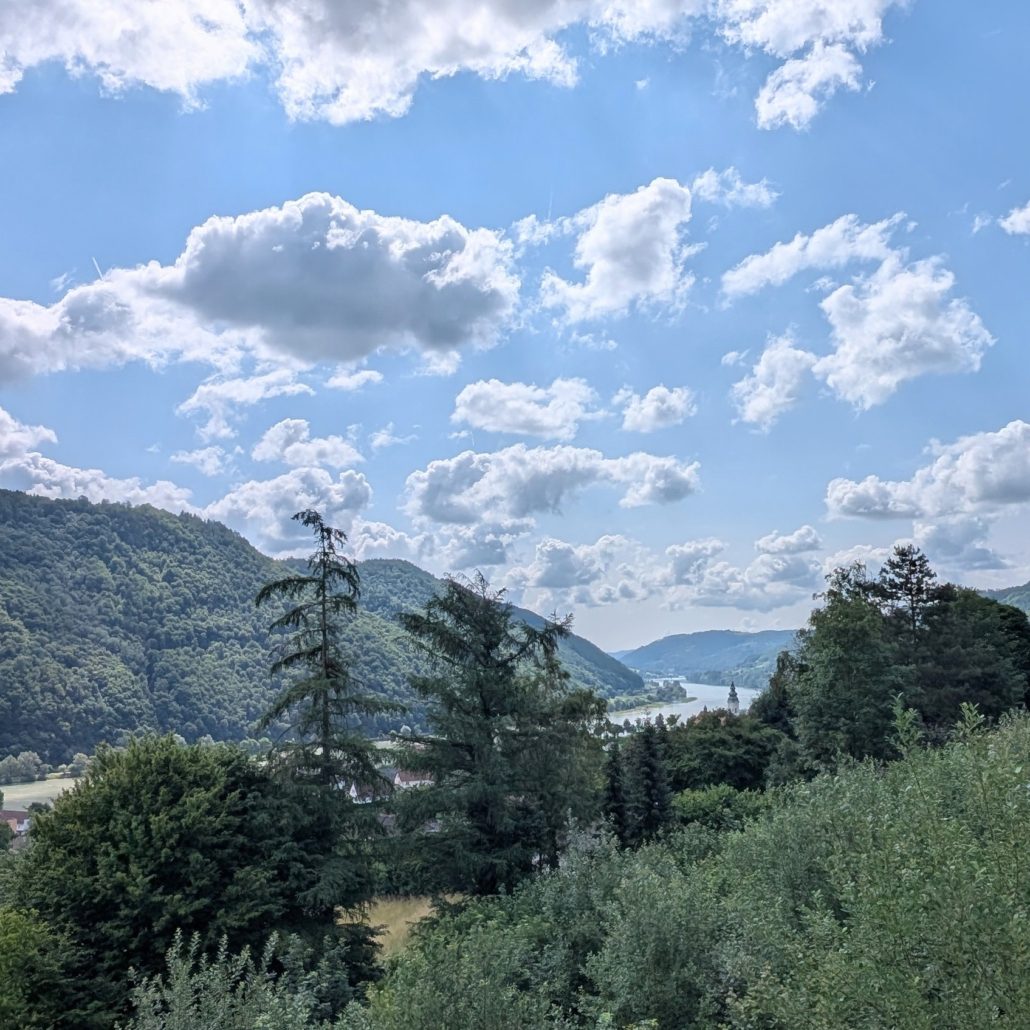 Waldbewachsene Hügel mit Blick auf ein Flusstal unter blauem Himmel mit weißen Cumuluswolken.