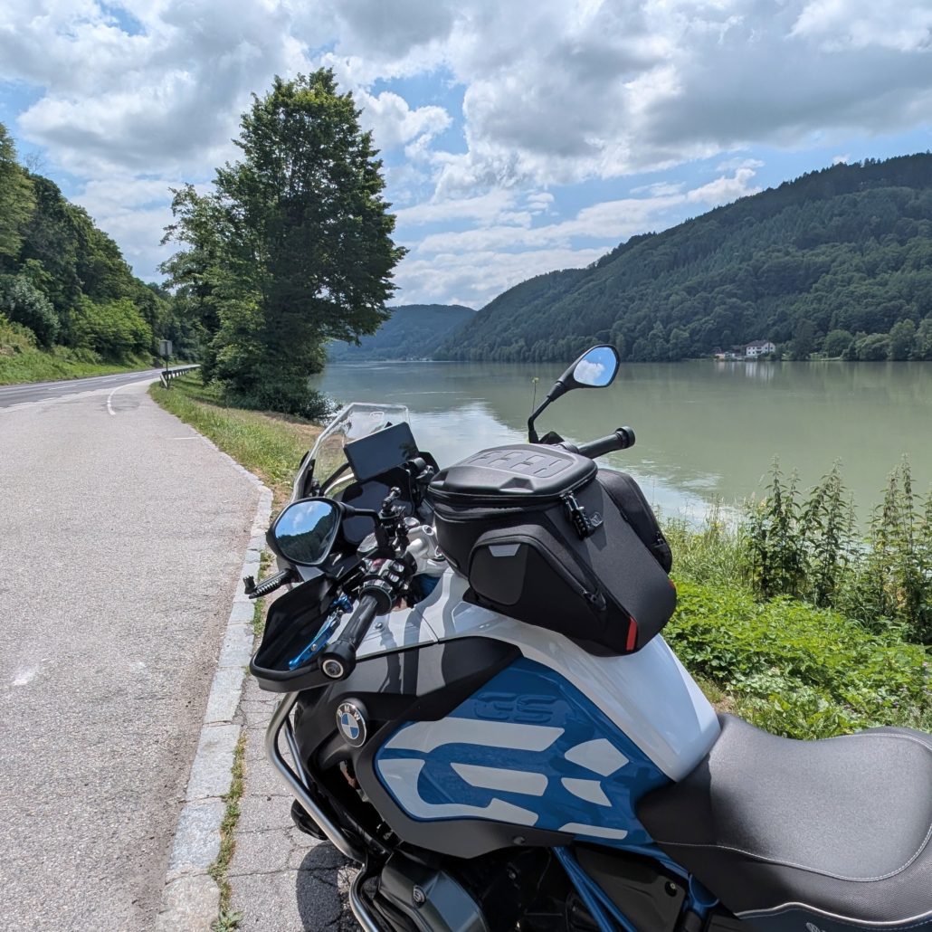 Detailansicht eines weiß-blauen Motorrads am Ufer der Donau, mit bewaldeten Hügeln im Hintergrund.