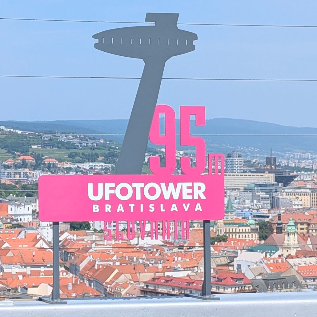 Schild "UFO TOWER BRATISLAVA" und "95m" vor der Dachlandschaft der Altstadt von Bratislava.