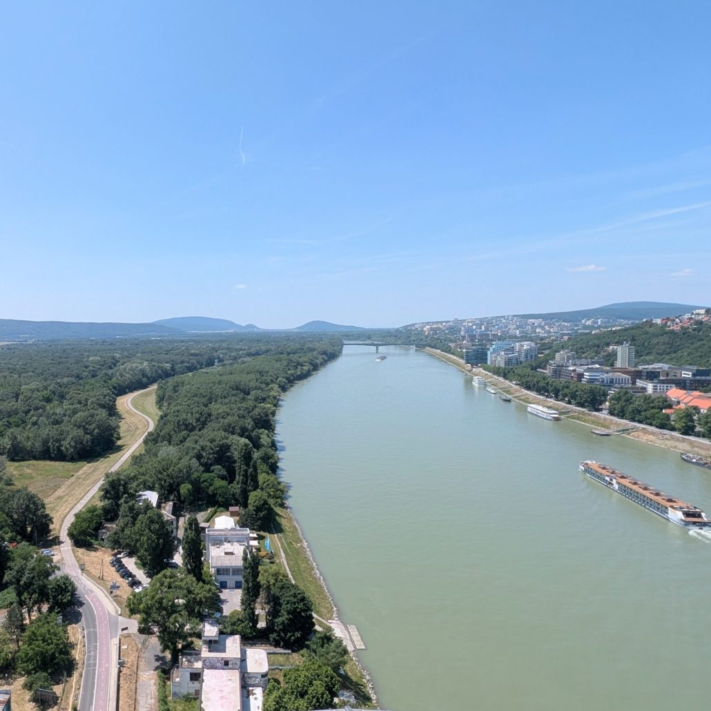 Panoramablick auf die Donau in Bratislava, mit Brücke, grünen Ufern und Stadtkulisse unter blauem Himmel.
