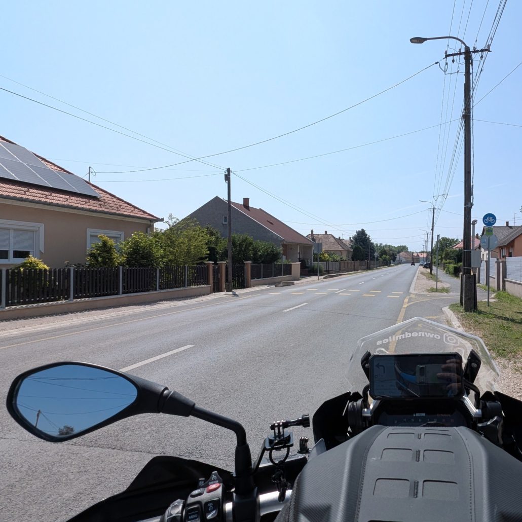Motorrad-Cockpit auf einer Dorfstraße, Häuser links, Laternenpfähle rechts.