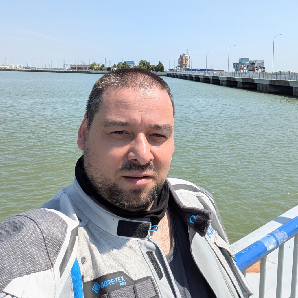 Selbstporträt eines Mannes in einer grau-blauen GORE-TEX Motorradjacke mit schwarzem Halstuch an einem Wasserkraftwerk.