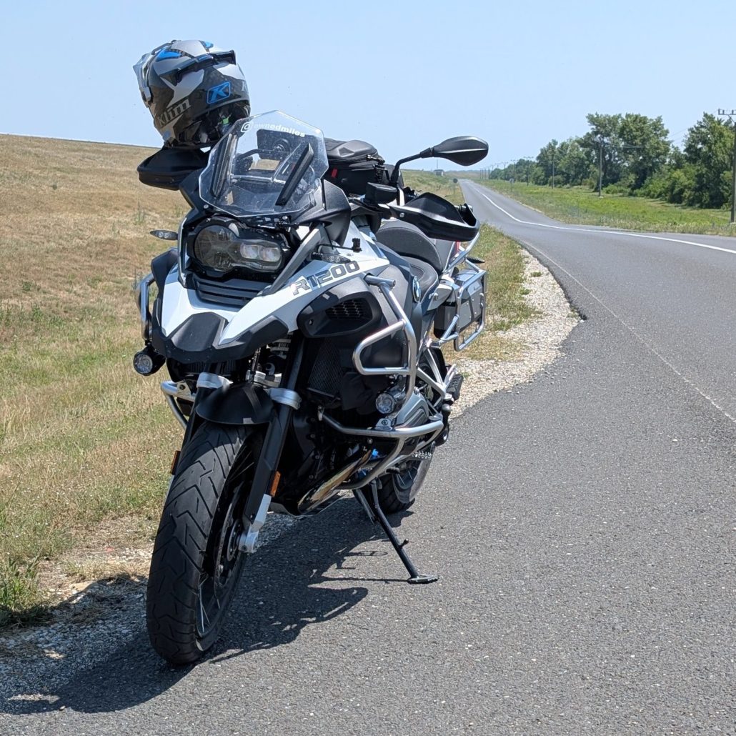Silber-schwarzes BMW R 1200 GS Motorrad mit einem Helm am Lenker und montierten Schutzbügeln an einer Landstraße unter blauem Himmel.