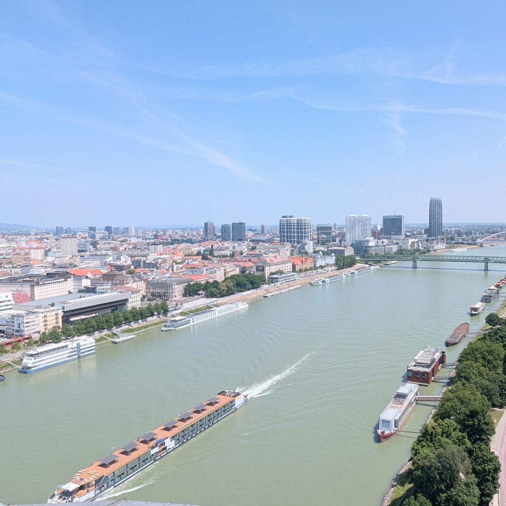 Blick über die Donau auf die Skyline von Bratislava mit Hochhäusern, Brücken und Flusskreuzfahrtschiffen bei Tag.