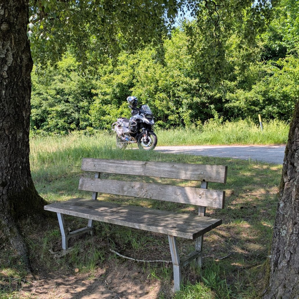 Holzbank im Schatten zweier Bäume, mit einem grauen BMW R 1200 GS Motorrad im sonnigen Hintergrund.