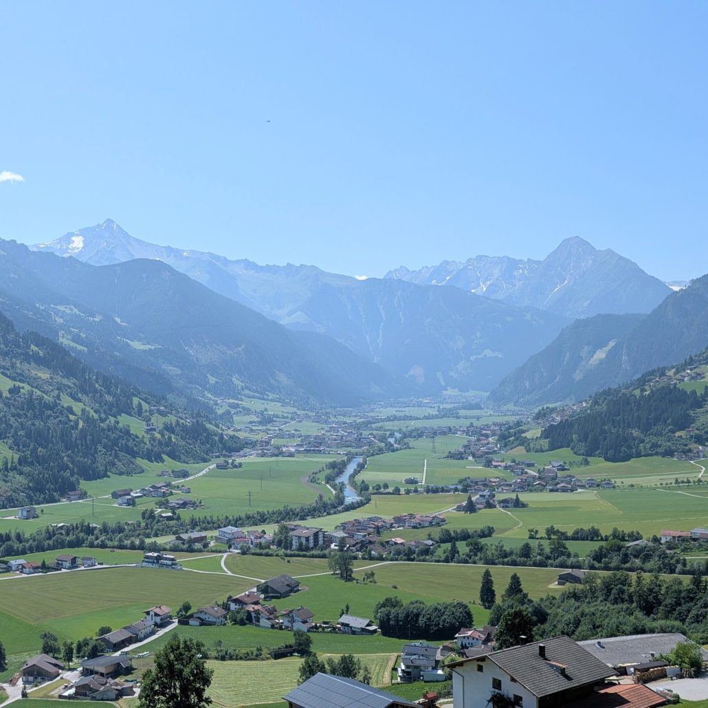 Motorrad-Sitzansicht eines tiefen, grünen Tals mit einem Flusslauf und einem Dorf, umgeben von schneebedeckten Alpengipfeln.