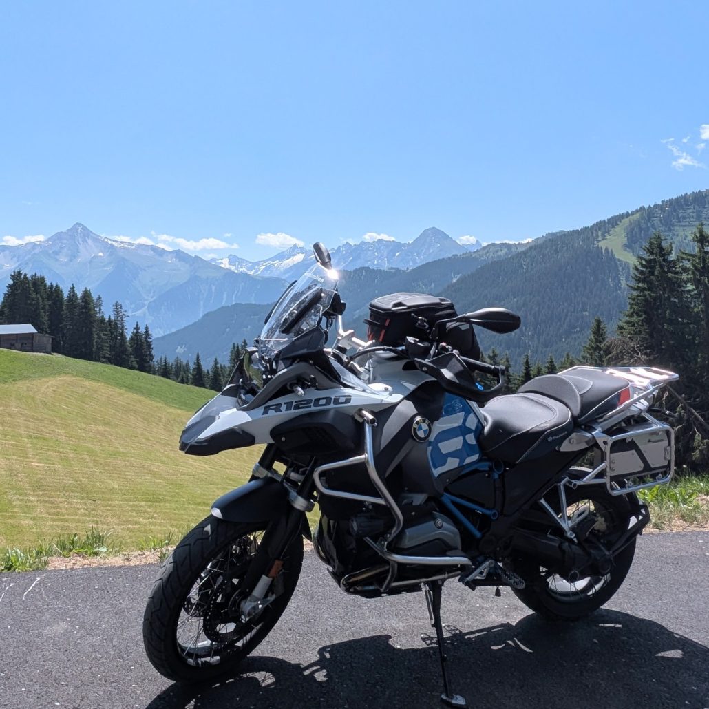 Helle BMW R 1200 GS Adventure Motorrad auf einer Bergstraße mit schneebedeckten Alpen im Hintergrund.