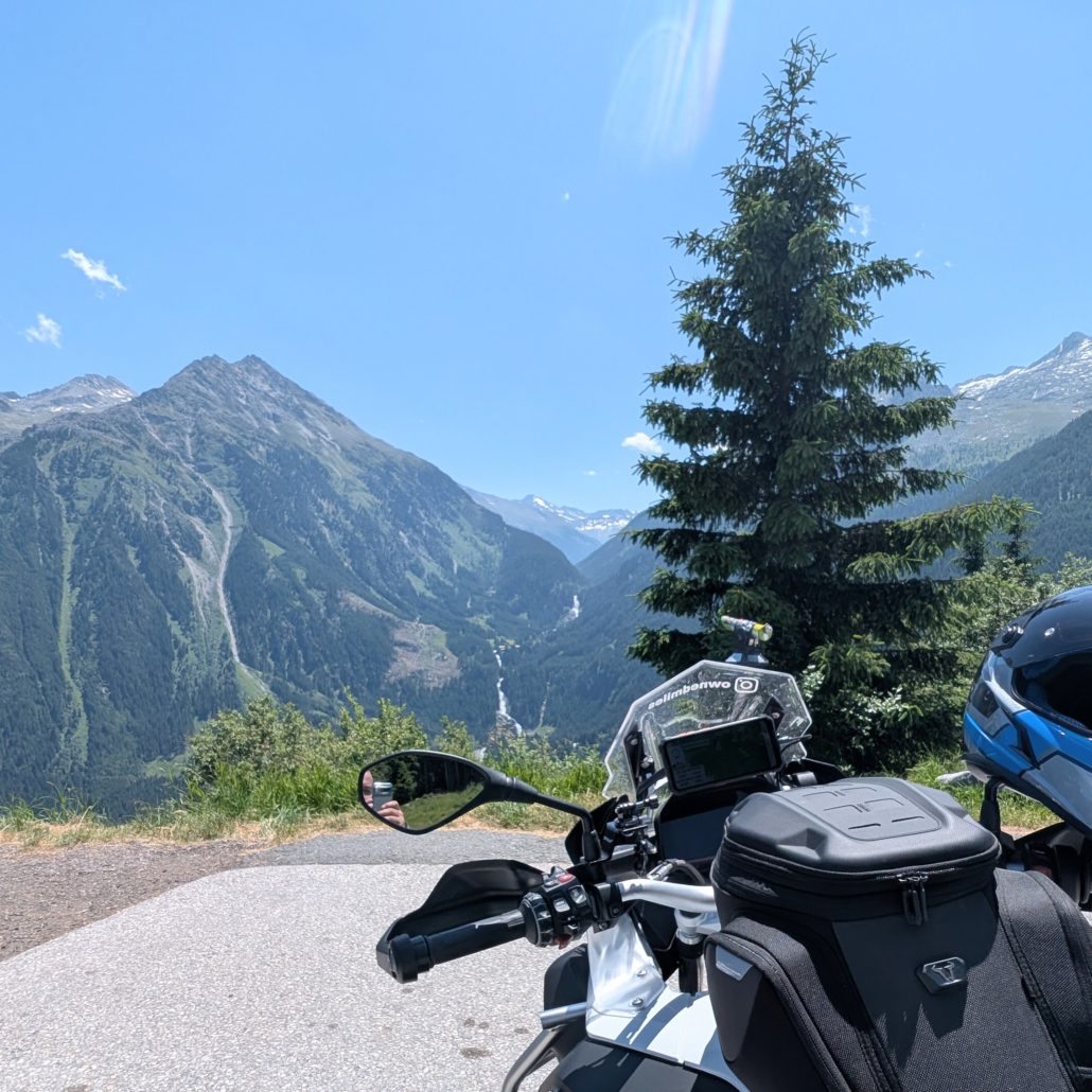 Aussicht von einer Bergstraße auf ein weißes Motorrad mit schwarzer Gepäcktasche und Helm vor einem alpinen Tal.