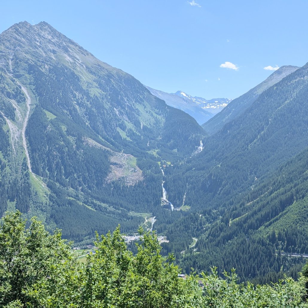 Panoramablick auf ein enges Alpental, umgeben von steilen, bewaldeten Hängen und einem mehrstufigen Wasserfall in der Mitte.
