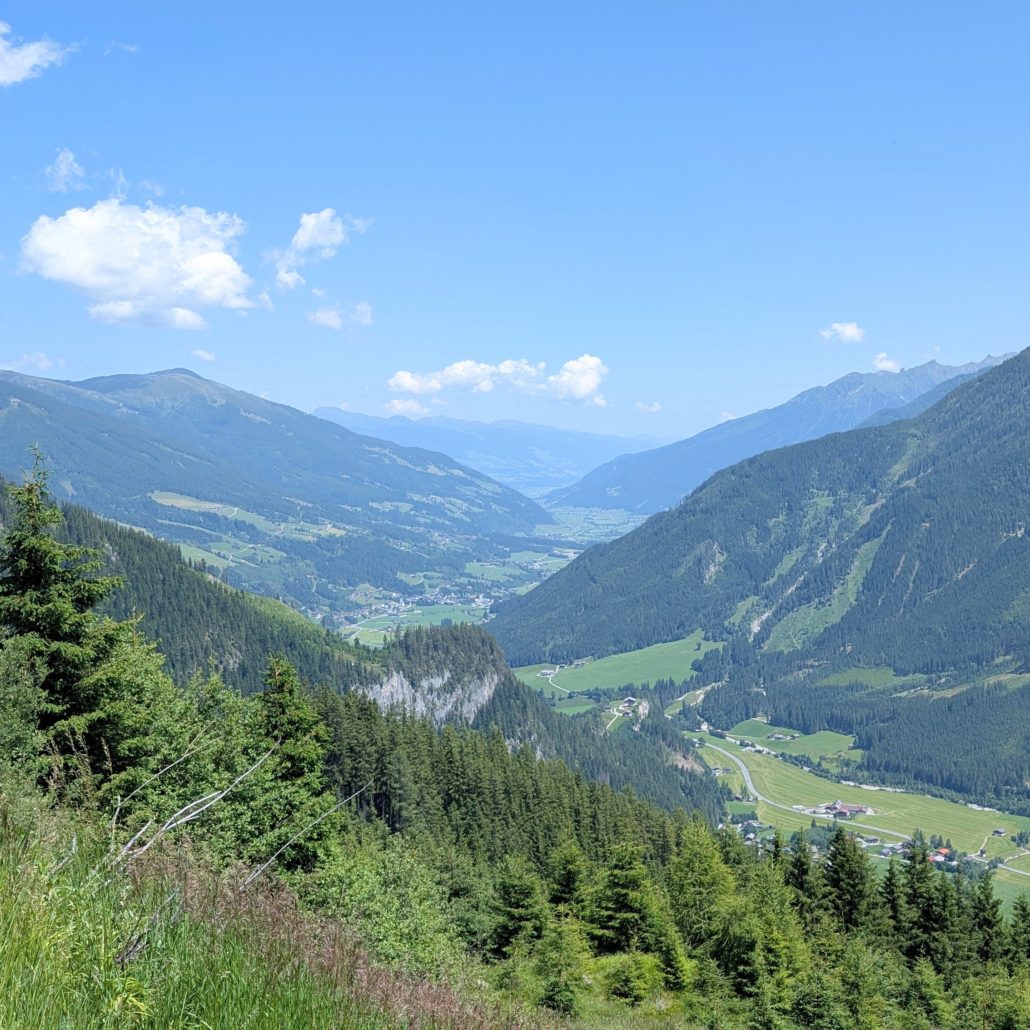 Weitwinkelaufnahme eines tiefen, grünen Alpentals, umgeben von bewaldeten Hängen unter blauem Himmel mit weißen Wolken.