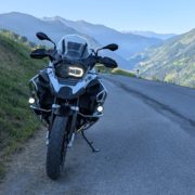 BMW F 1200 GS Adventure Motorrad in Weiß und Schwarz auf einer Bergstraße mit Alpenpanorama im Gegenlicht.