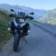 BMW F 1200 GS Adventure Motorrad in Weiß und Schwarz auf einer Bergstraße mit Alpenpanorama im Gegenlicht.