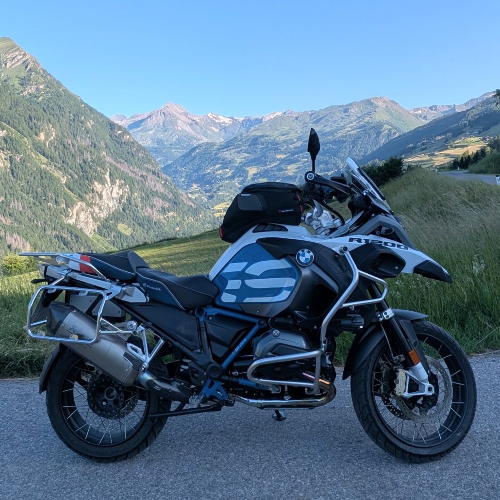 Gerade asphaltierte Straße führt in ein Tal mit grünen Berghängen, Schneebedeckten Gipfeln und klarem blauem Himmel.