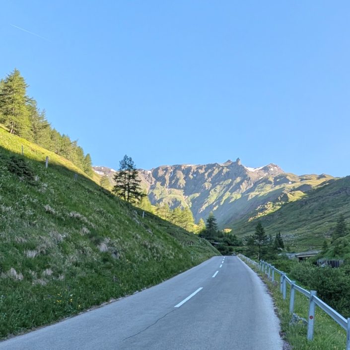 Asphaltierte Bergstraße mit weißer Mittellinie führt durch ein grünes Tal, umgeben von steilen Hängen und Gipfeln.