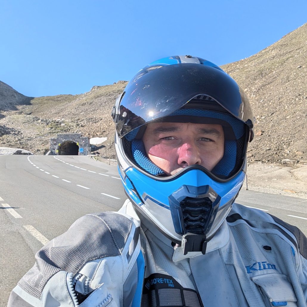 Motorradfahrer in blau-weißem Helm und KLIM GORE-TEX Jacke auf einer Bergstraße mit Tunnel im Hintergrund.