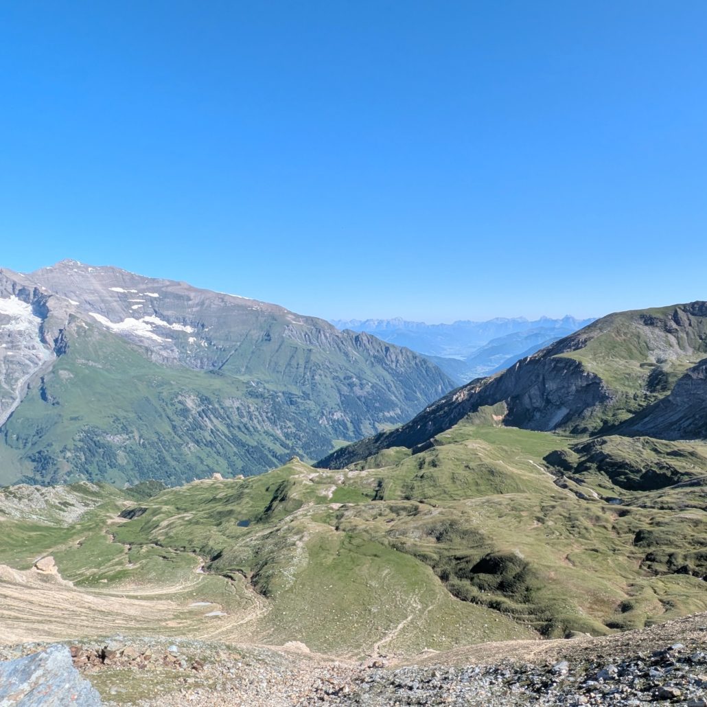 Grüne, hügelige Alpenlandschaft mit Serpentinenpfaden und Blick auf ferne, schneebedeckte Gipfel unter klarem Himmel.