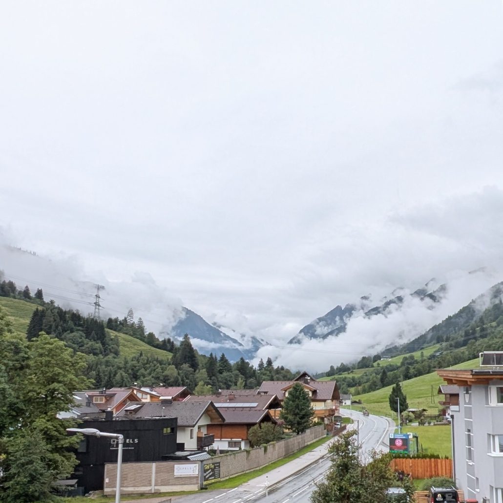 Dorfansicht in einem bewölkten alpinen Tal mit bewaldeten Hängen und nebelverhangenen Gipfeln, Häuser entlang einer Straße.