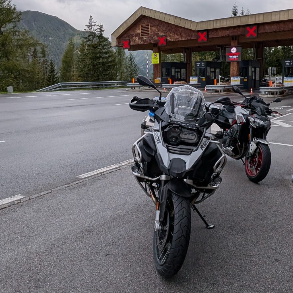Zwei Motorräder, eine weiße BMW R 1200 GS und eine rote Sportmaschine, stehen vor einer Mautstation in den Bergen.