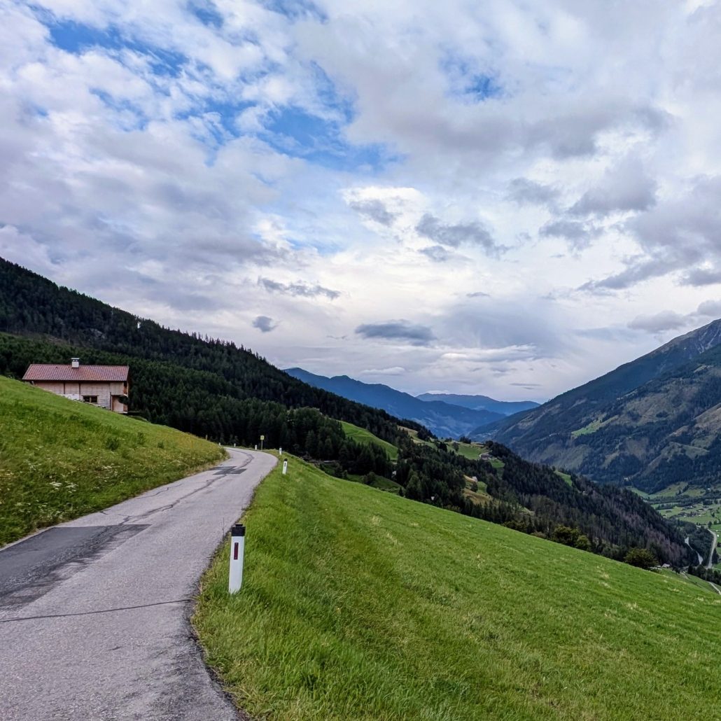 Eine kurvige Asphaltstraße führt einen grünen Berghang hinauf, mit einem braunen Haus links und einer bewaldeten Schlucht rechts.