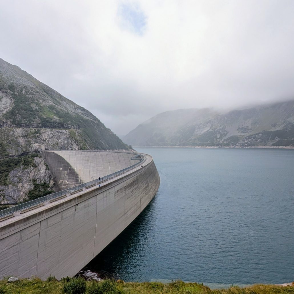 Ein breiter Alpensee umgeben von dicht bewaldeten Berghängen und einer Staumauer.