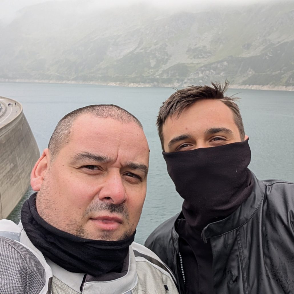 Zwei Motorradfahrer, einer davon mit schwarzem Gesichts-Tuch, machen ein Selfie vor einer hohen Staumauer und einem Bergsee.