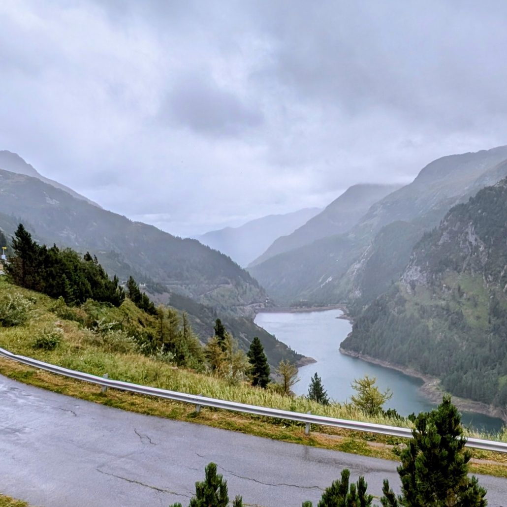 Ein stahlblauer Stausee tief in einem grünen, nebelverhangenen Gebirgstal, gesehen von einer Serpentinenstraße.