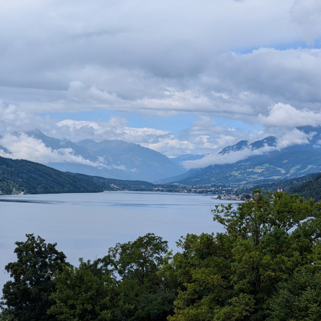 Ein See, umgeben von dichten grünen Bäumen und einer Bergkette, deren Gipfel teilweise von Wolken bedeckt sind.