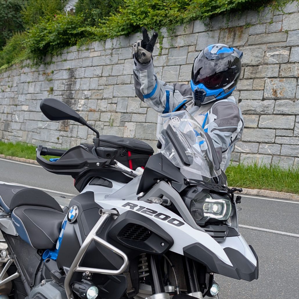 Ein Motorradfahrer in grau-blauer Montur und Helm steht neben einem weißen BMW R 1200 Adventure-Motorrad und macht ein V-Zeichen.
