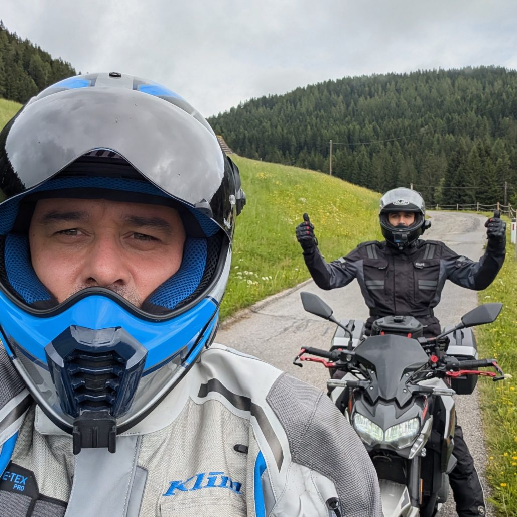 Zwei Motorradfahrer in voller Schutzkleidung auf einer schmalen Bergstraße; der vordere macht ein Selfie.