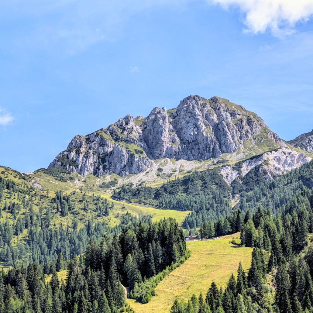 Eine zerklüftete, graue Felsformation dominiert die Berglandschaft, umgeben von einem Nadelwald und grünen Almwiesen.