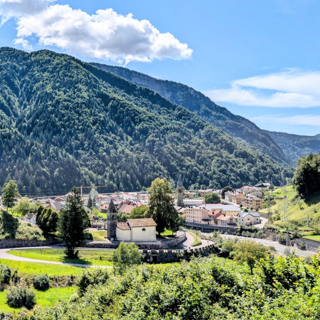 Idyllisches Bergdorf im Tal, umgeben von dicht bewaldeten, steilen Hängen unter blauem Himmel mit weißen Wolken.