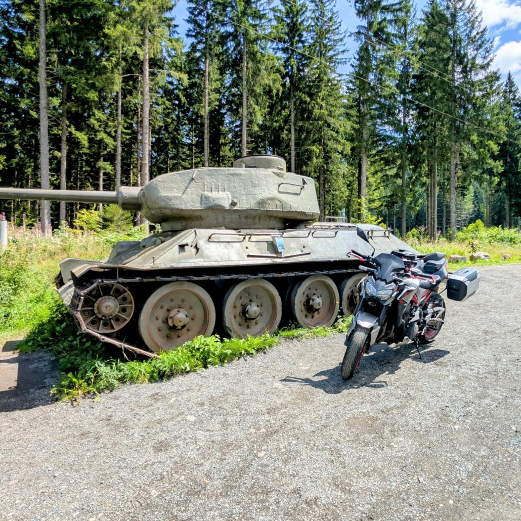T-34/85 Panzerdenkmal in einem Waldgebiet, daneben ein modernes graues Motorrad auf einem Schotterweg.