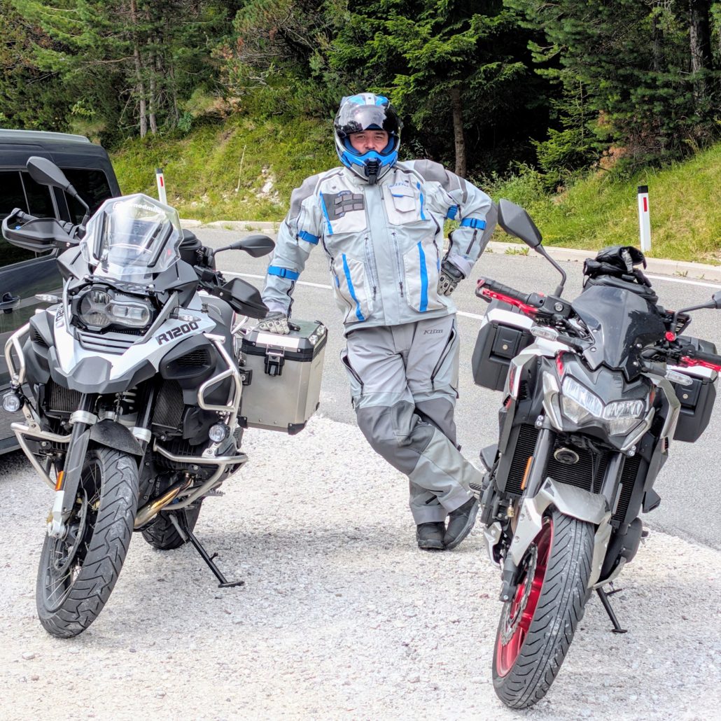 Motorradfahrer in grauer und blauer Motorradkleidung steht zwischen einer weißen BMW R1200 GS und einem silbernen Naked Bike.