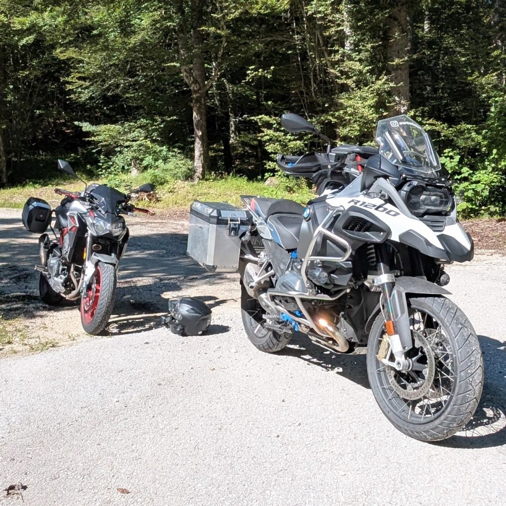 Zwei Motorräder parken an einem Waldrand auf Schotter: eine weiße BMW R1200 GS Adventure mit Koffern und ein rotes Naked Bike.