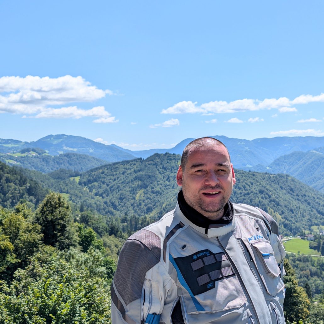 Motorradfahrer in grauer Adventure-Jacke (GORE-TEX) lächelt vor einer weiten grünen Berglandschaft mit blauem Himmel.