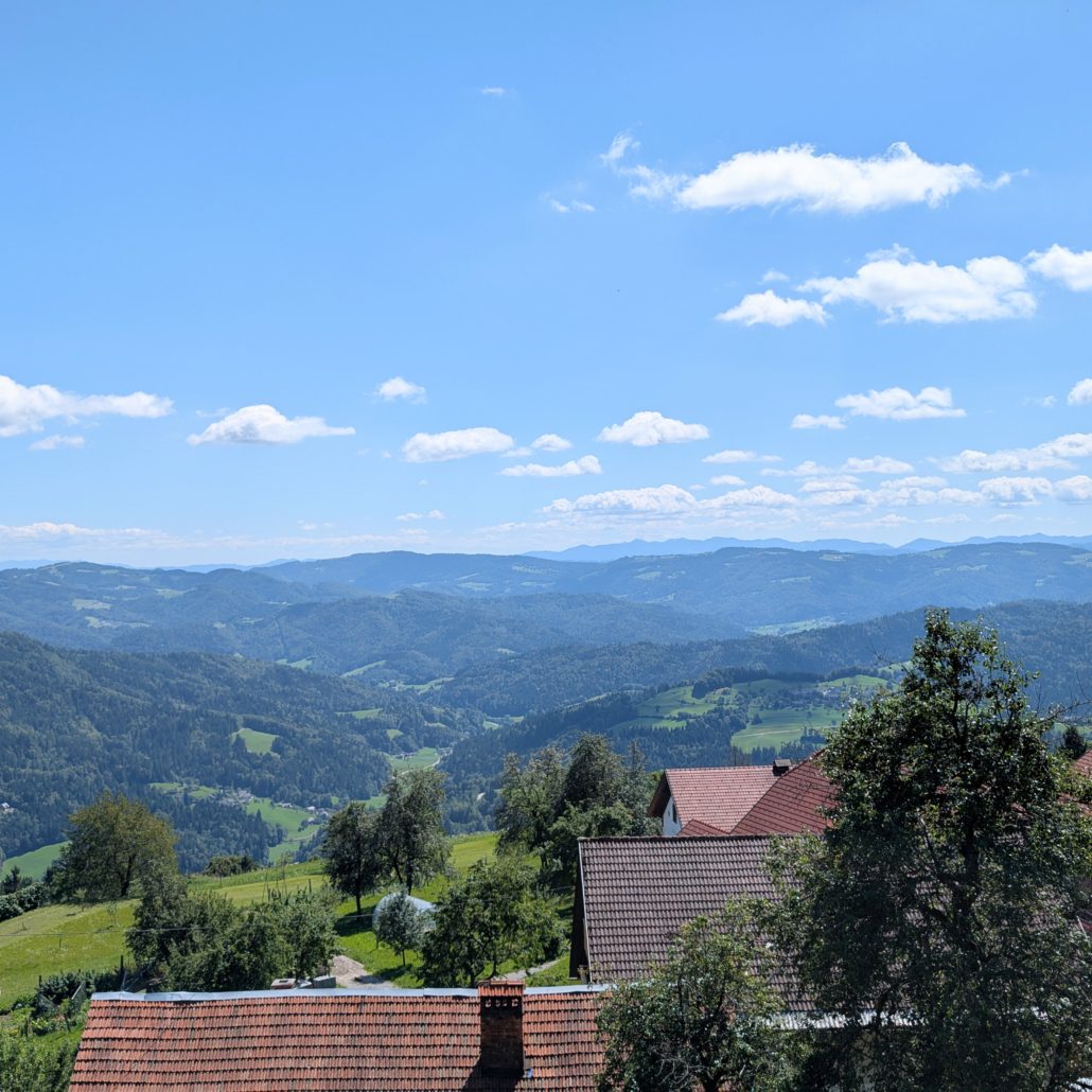 Blick auf sanfte, bewaldete Hügel unter blauem Himmel mit weißen Wolken; rote Ziegeldächer im Vordergrund.