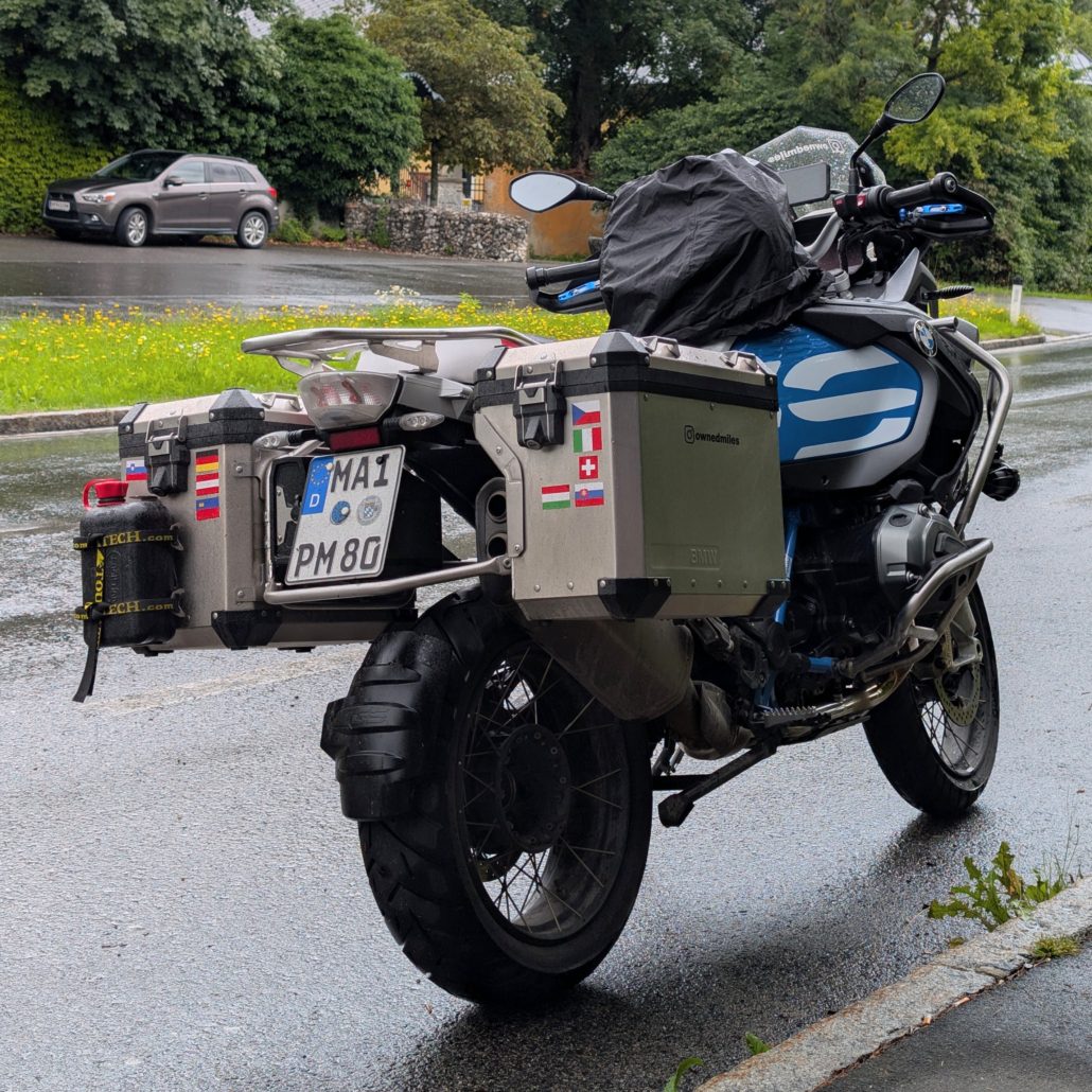 Motorrad (BMW R 1200 GS) auf nasser Straße, von hinten mit silbernen Seitenkoffern und Länder-Aufklebern.