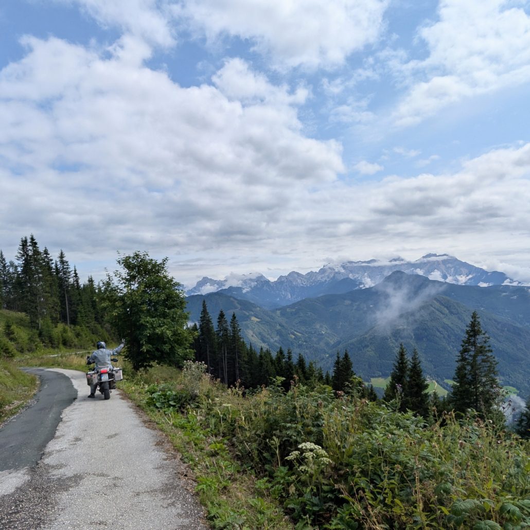 Motorradfahrer auf einem Asphaltweg mit Bergblick auf hohe, teils schneebedeckte Gipfel unter bewölktem Himmel.