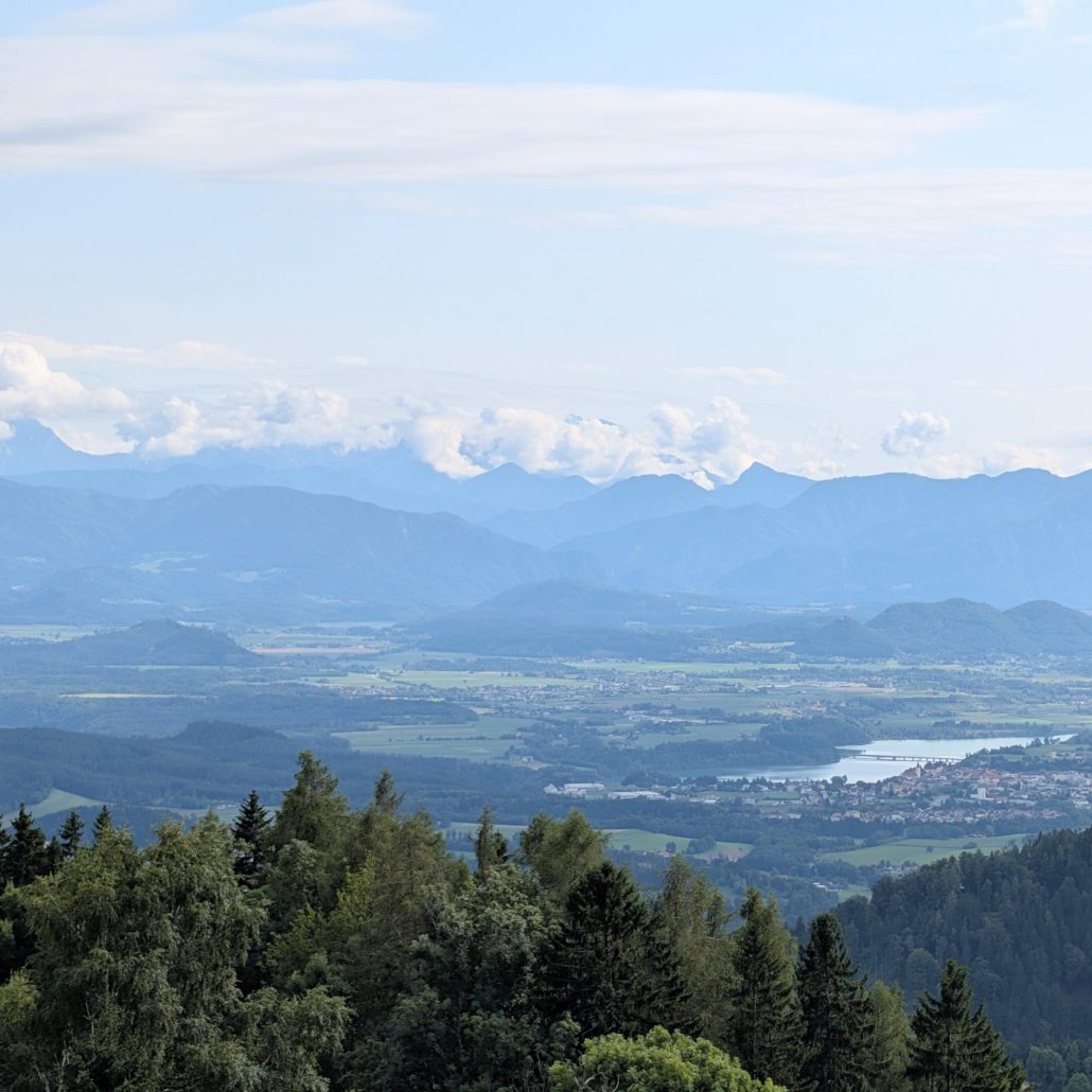 Weitblick über bewaldete Hügel, ein Tal mit See und Stadt, bis zu einer Kette blauer Alpenberge am Horizont.