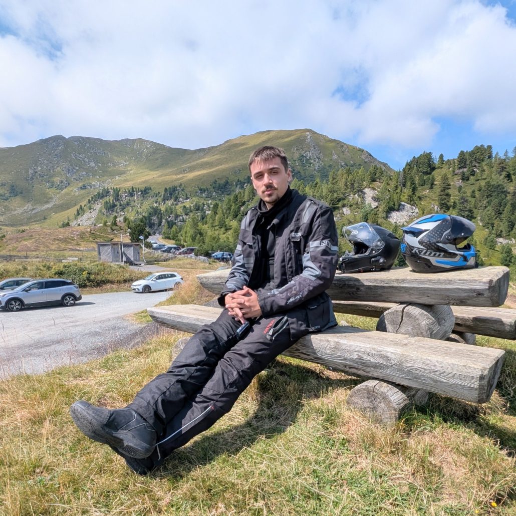 Motorradfahrer in schwarzer Abenteuerausrüstung sitzt mit zwei Helmen auf einer Holzbank vor Bergpanorama.