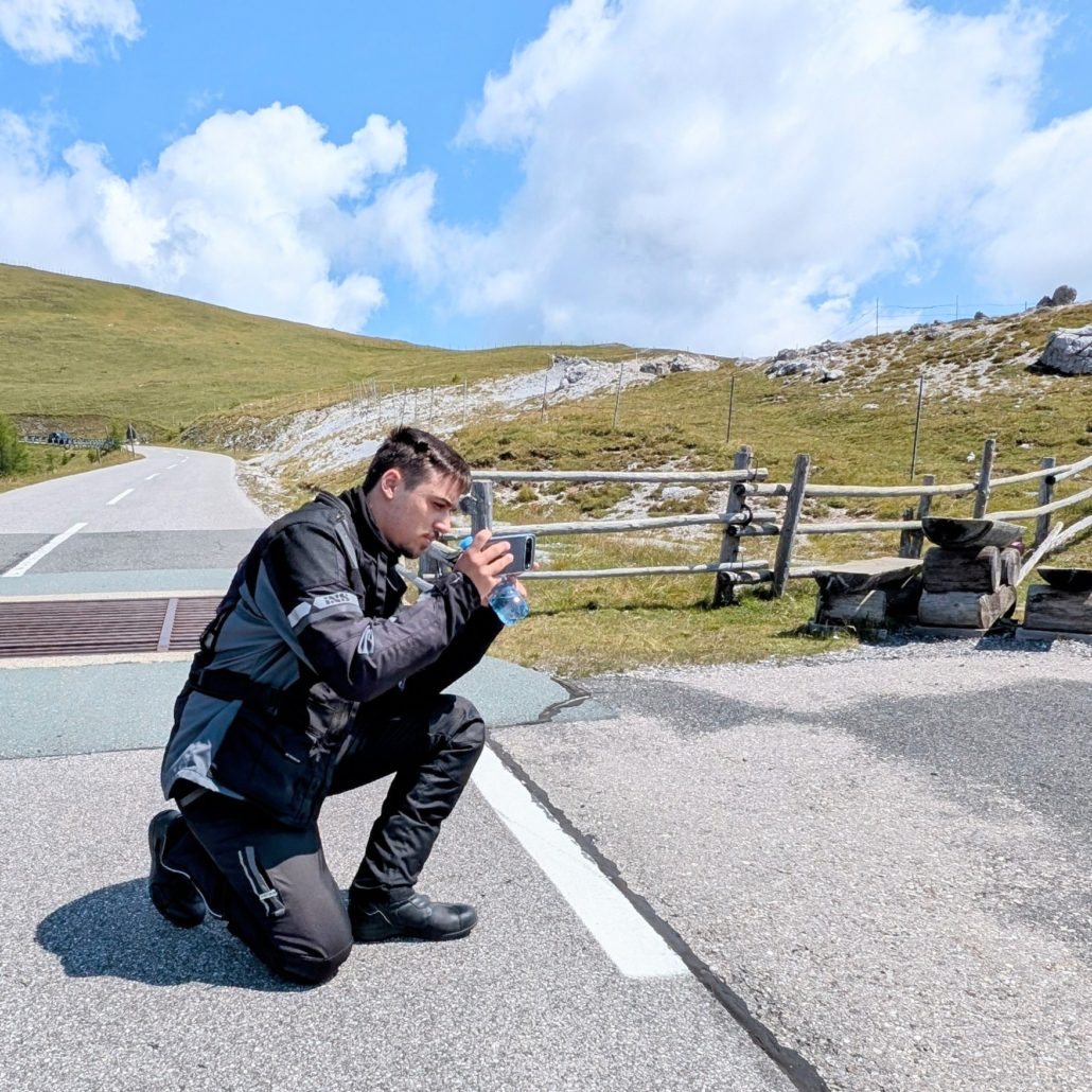 Ein Motorradfahrer in schwarzer Motorrad-Kombination kniet auf einer Passstraße und fotografiert mit seinem Smartphone.